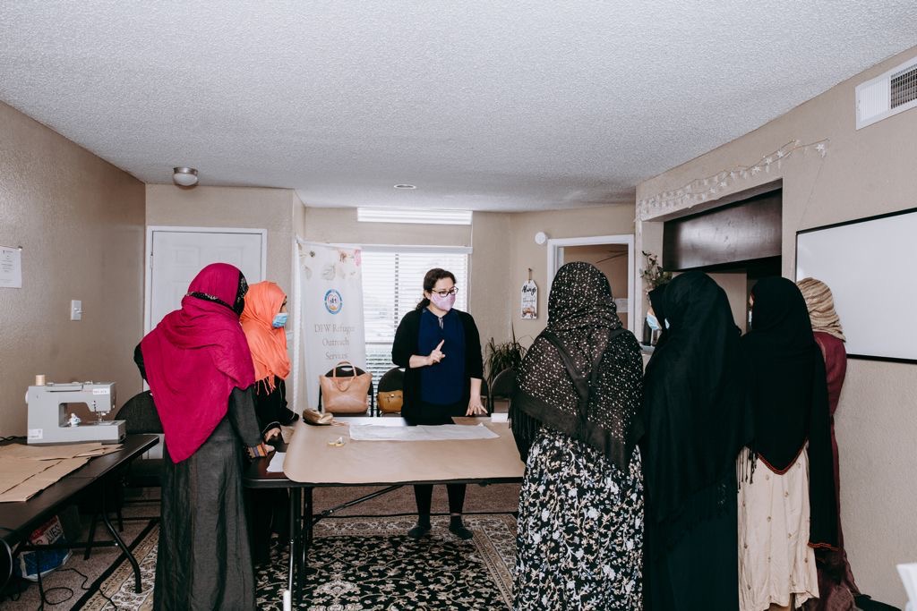Refugee women gathered around a sewing instructor during a Hope Boutique class &mdash; a sewing machine and DFWROS banner visible in the background