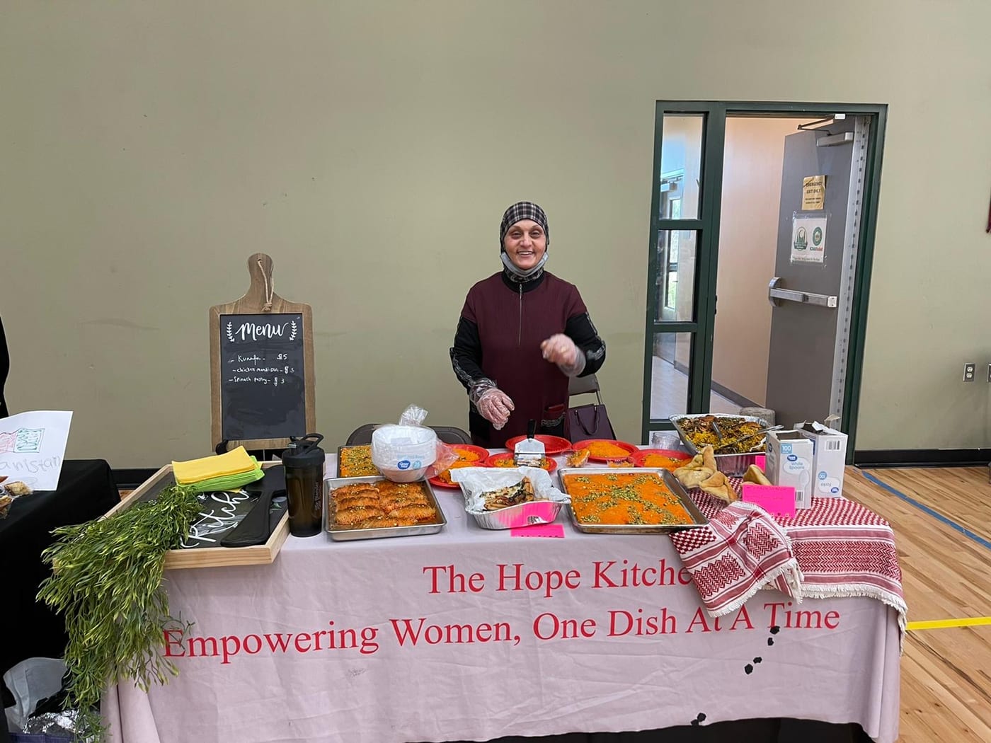 A Hope Kitchen chef at her stall, behind a banner reading &lsquo;The Hope Kitchen &mdash; Empowering Women, One Dish At A Time&rsquo;